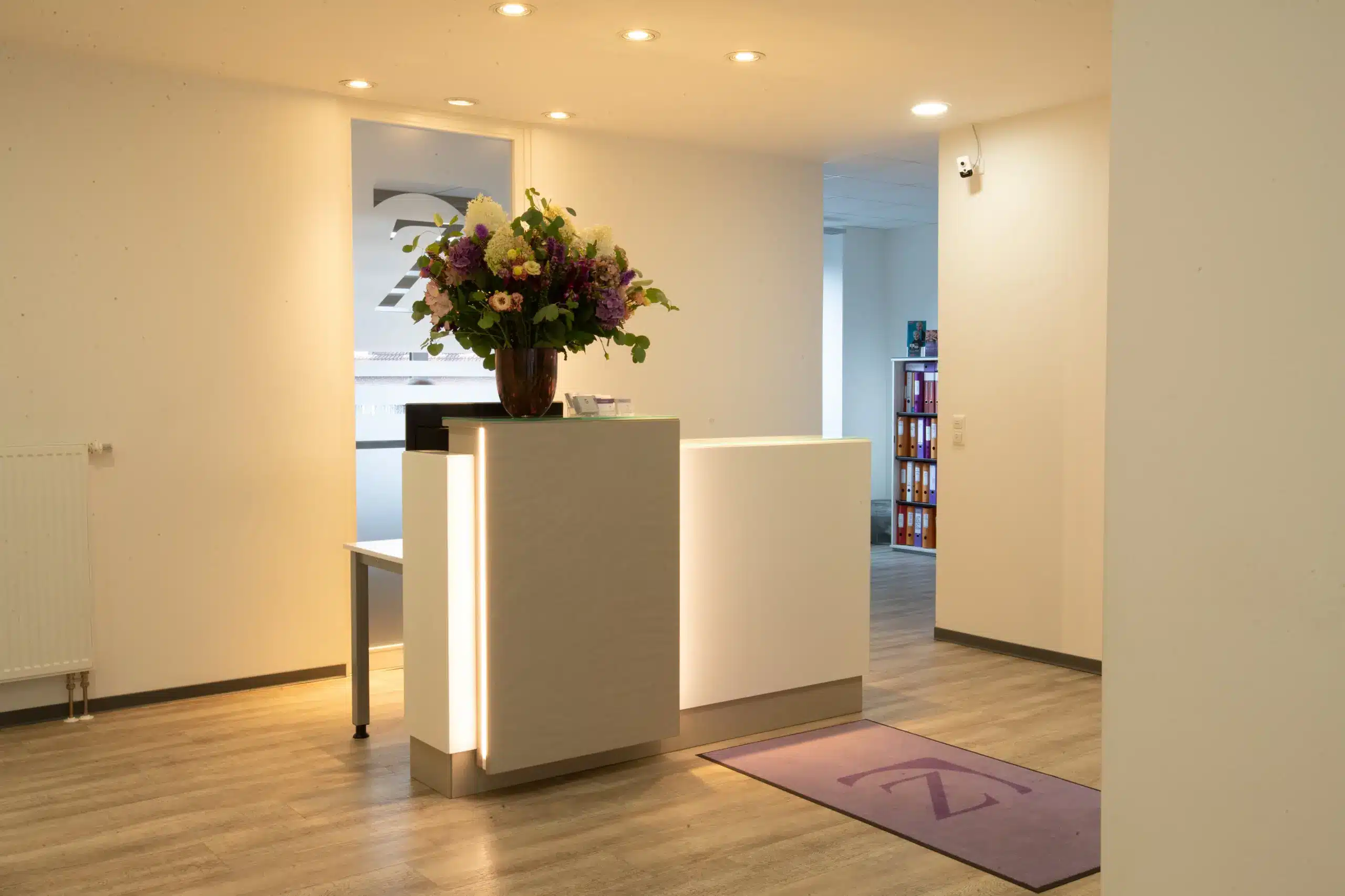 Warm beleuchteter Empfangstresen der Zahnarztpraxis Dr. Menninger in Pinneberg mit Blumenstrauß und Fußmatte mit Praxislogo